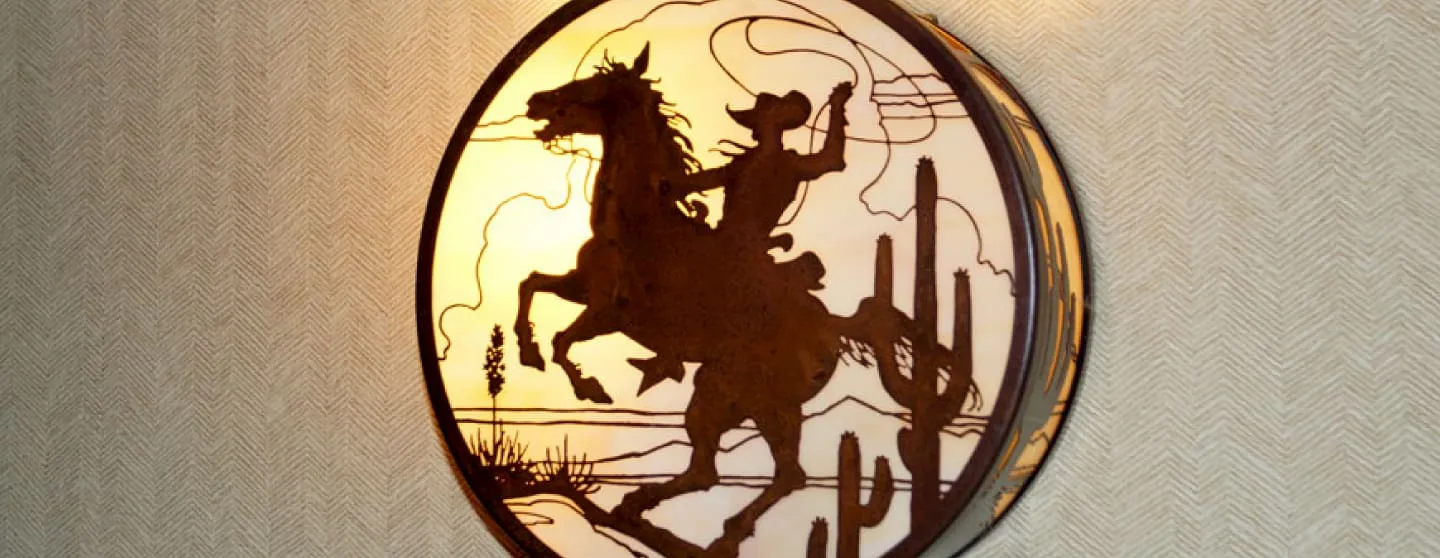 Round wall light fixture with a silhouette of a cowboy on a rearing horse and desert cacti background.