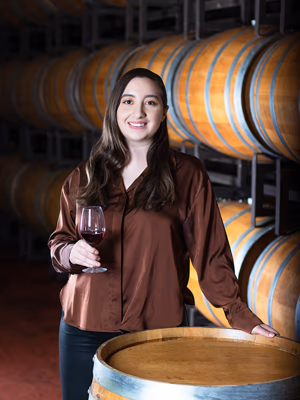 Andrea Zermeno standing in a barrel room holding a glass of red wine and smiling