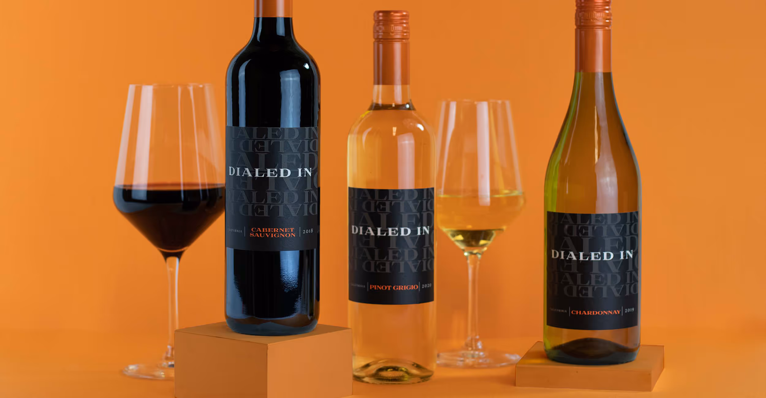 A trio of Dialed In wine bottles next to wine glasses over an orange background