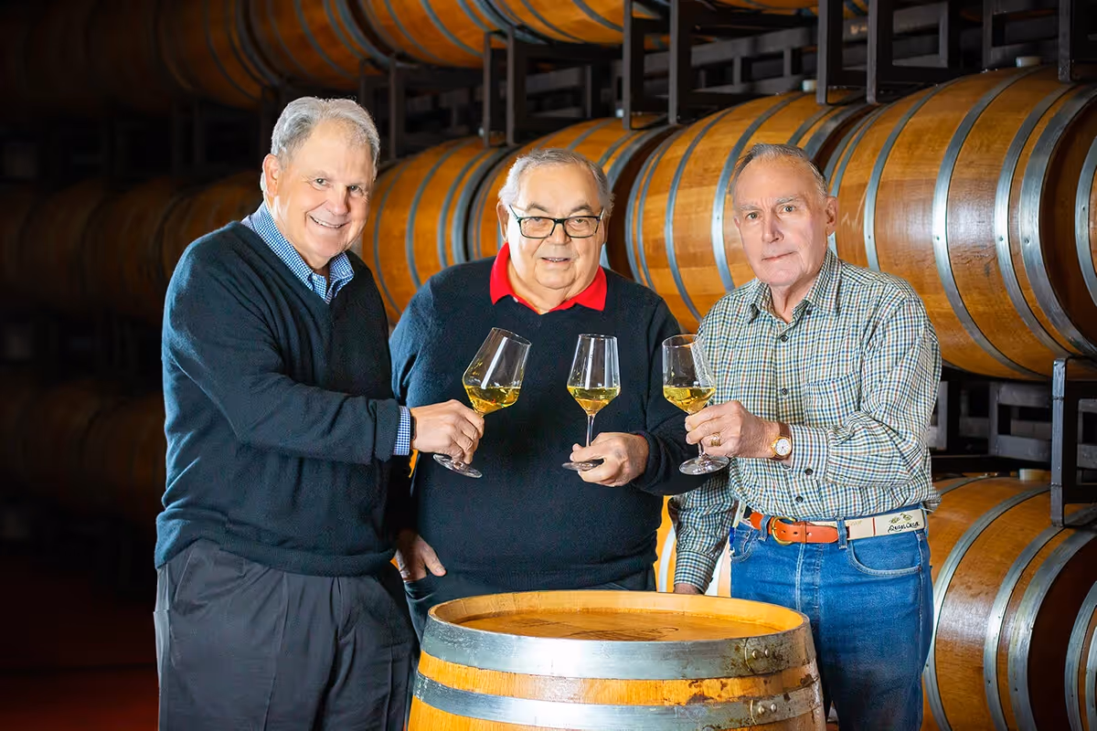Joseph, Fred, and John Franzia pose in the barrel room each holding a glass of chardonnay in 2022