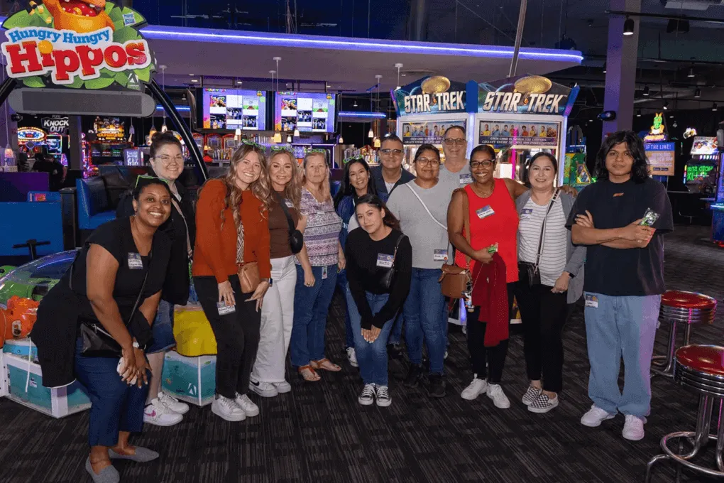 A group of people poses together smiling in an arcade setting, surrounded by social walls and colorful games, including Hungry Hungry Hippos and Star Trek-themed machines.