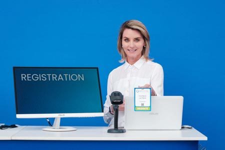  smiling woman at a registration desk holds up a printed delegate badge beside a computer screen that reads registration