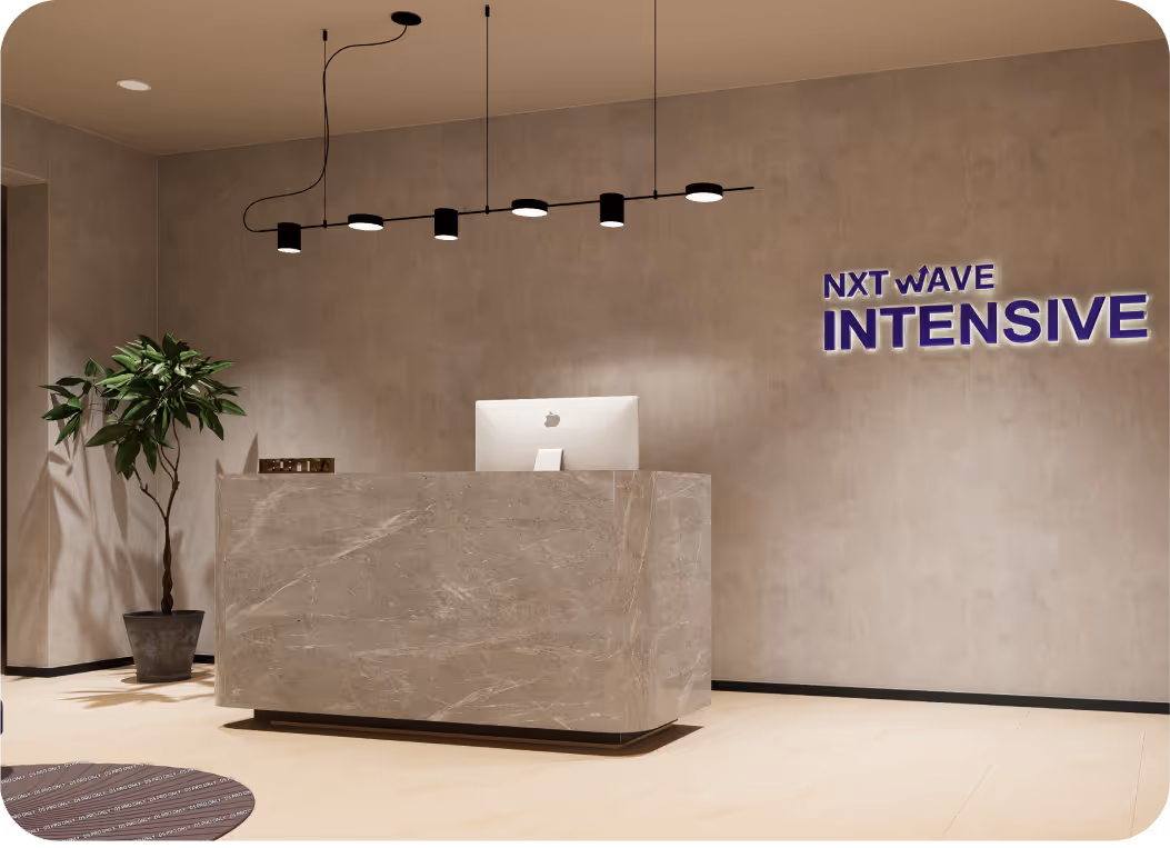 Modern reception area with a marble desk, an Apple computer, a potted plant, pendant lights, and the text 'NXT WAVE INTENSIVE' on the wall.
