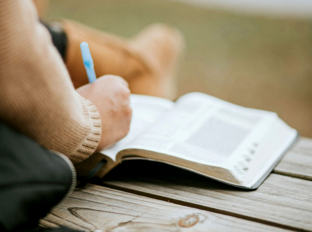 Person in a sweater writing in a Bible with a pen outdoors on wooden surface.