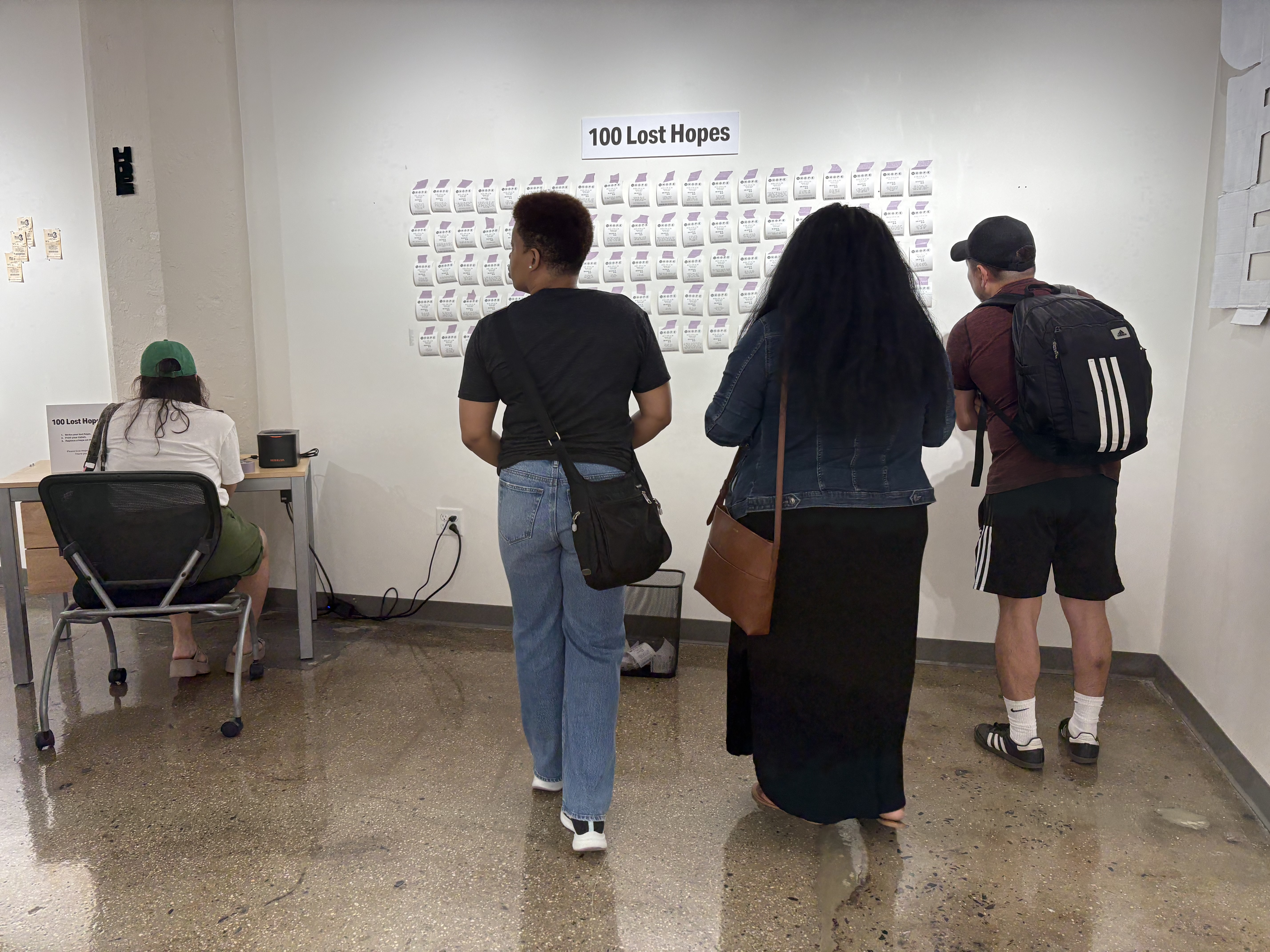 Viewers looking at the work, one using a computer. On the wall is a sign that says "100 Lost Hopes." Underneath it are 100 fake lottery tickets taped to the wall.