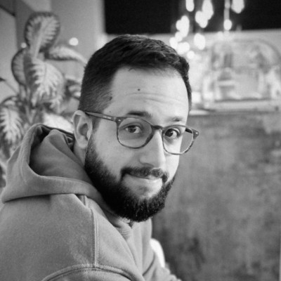 Man with glasses and a beard wearing a hoodie, looking over his shoulder and smiling in a cozy indoor setting with soft lighting and plants in the background, black and white photo