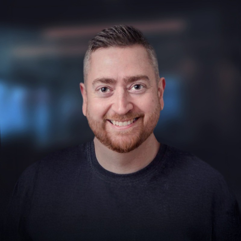 Smiling man with short hair and a beard wearing a dark shirt, posed in front of a blurred indoor background