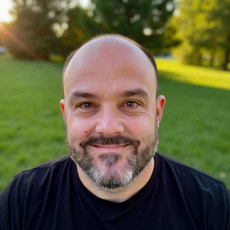 Smiling man with a beard and black shirt sitting outdoors on a grassy field with trees and sunlight in the background