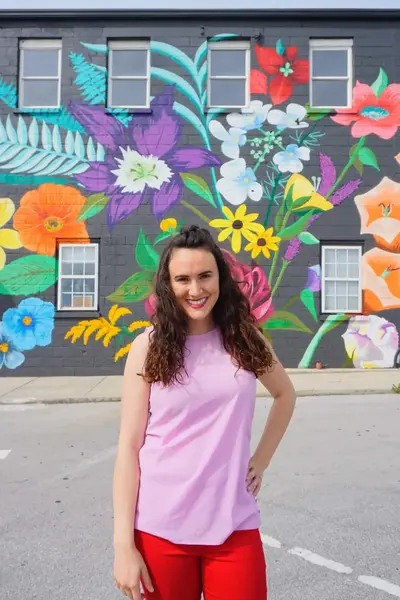 Smiling woman with curly hair wearing a pink sleeveless top and red pants stands in front of a colorful floral mural on a building wall.