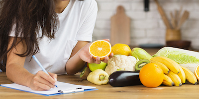 woman charting notes holding an orange on a table with fruits and vegetables