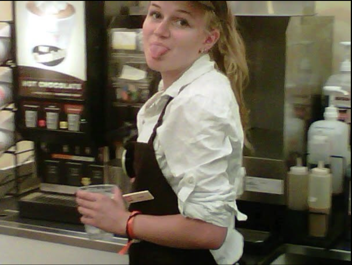 a young woman wearing a white button down and brown apron sticking out her tongue holding a clear cup. 