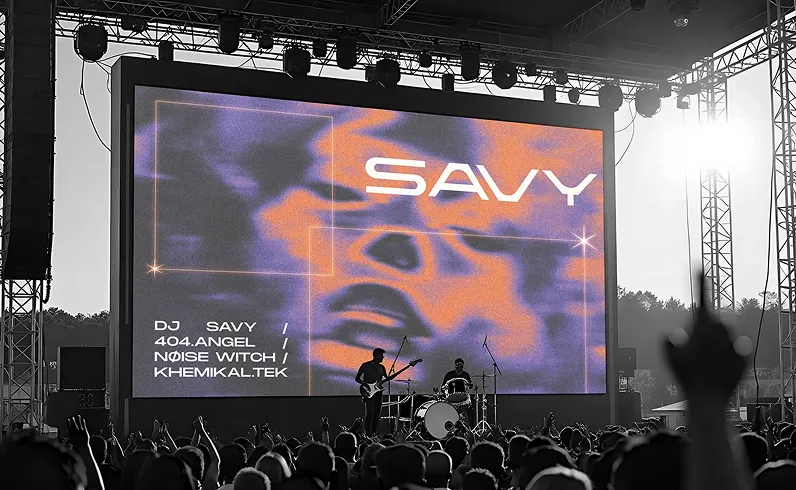 Black and white photo of a live music concert with a large screen displaying the word 'SAVY' and a list of performers: DJ Savy, 404.Angel, Noise Witch, and Khemikal.Tek, with a guitarist and drummer on stage in front of an audience.