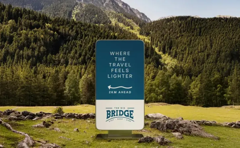 Sign in a grassy area with trees saying 'Where the travel feels lighter, 2km ahead, The Big Bridge'.