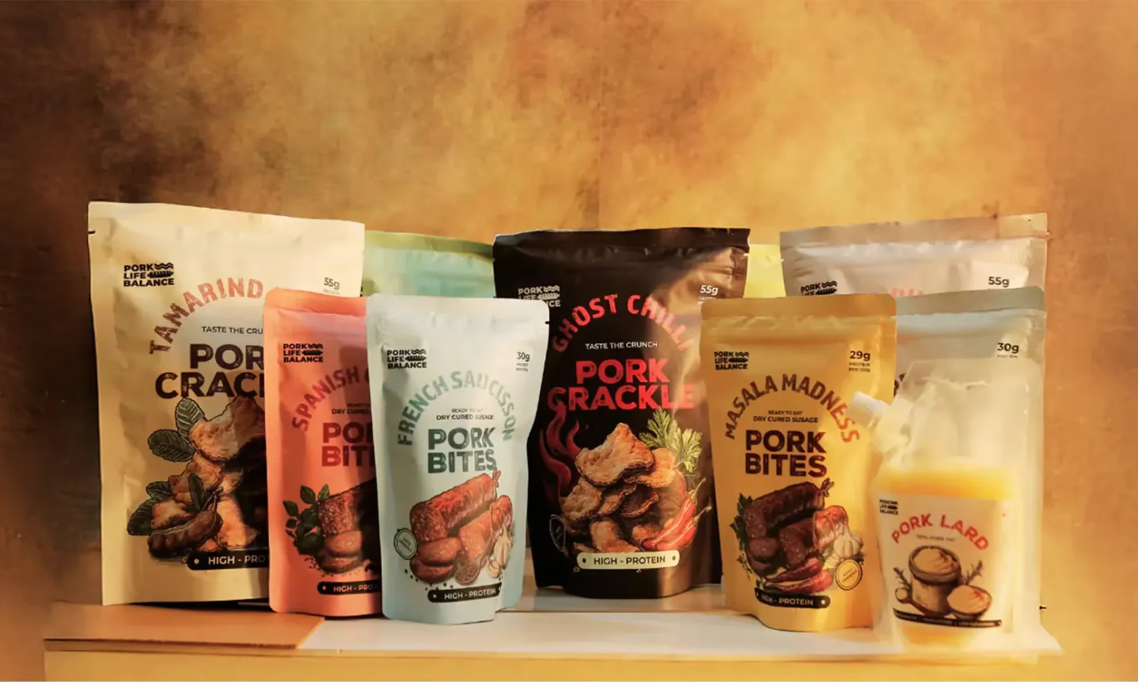 Seven colorful pouches of pork snacks and pork lard from Pork Life Balance, including tamarind pork crackles, Spanish pork bites, French sausage pork bites, ghost chili pork crackle, masa madness pork bites, and pork lard, arranged on a wooden surface with a warm brown background.