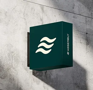 Square dark green sign mounted on a concrete wall with a white abstract logo of three wavy lines and the word 'ASSEMBLY' in capital letters on the side.