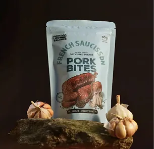 Package of Pork Bites dry cured sausage with fresh sauce seasoning, displayed on a wooden surface with garlic bulbs around it against a dark background.