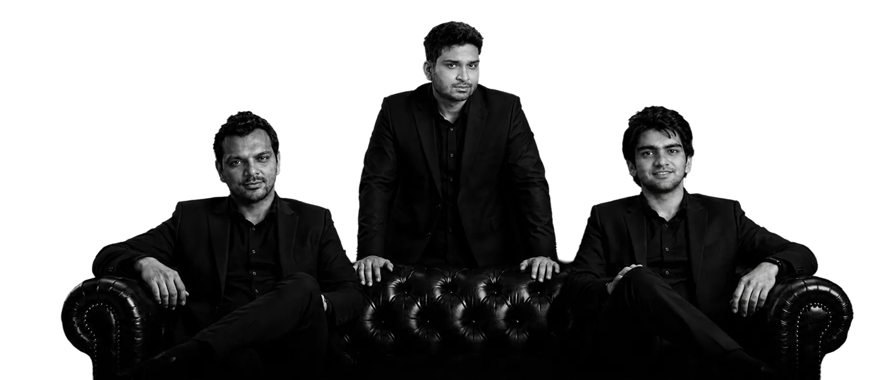 Black and white photo of three men in black suits: two seated on a tufted leather couch with arms on the couch, and one standing behind the couch with hands resting on its back.