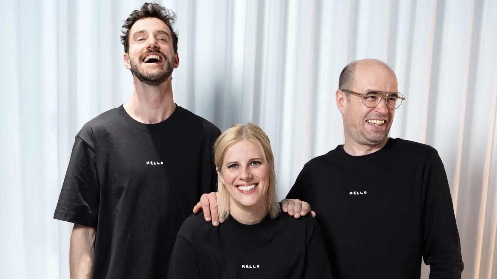 Drei fröhliche Teammitglieder posieren lachend in einheitlichen „HELLO“-Shirts vor einem hellen Hintergrund.