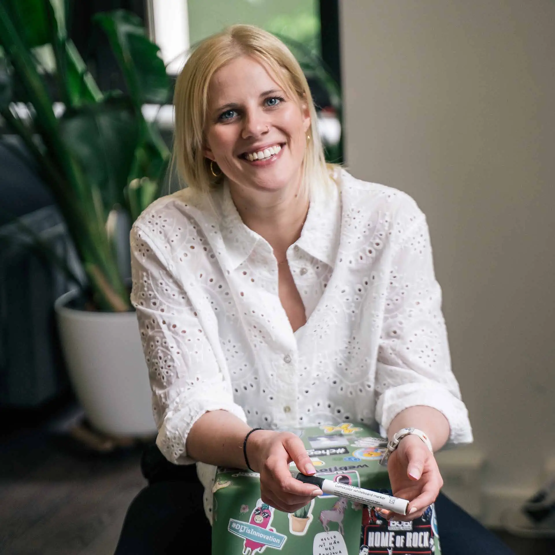 Kim Berger von HelloAgile. sie trägt ein schwarzen T-Shirt au dem "Be Nice, Say Hello" Steht. Sie steht vor einem blauen Hintergrund mit weißen Icons