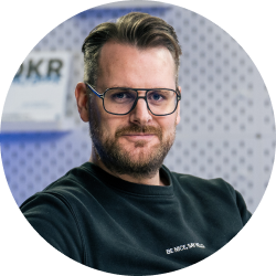 Ein Mann mit kurzen braunen Haaren, Bart und Brille trägt einen schwarzen Pullover vor einem unscharfen Hintergrund mit hellgrauem Muster.
