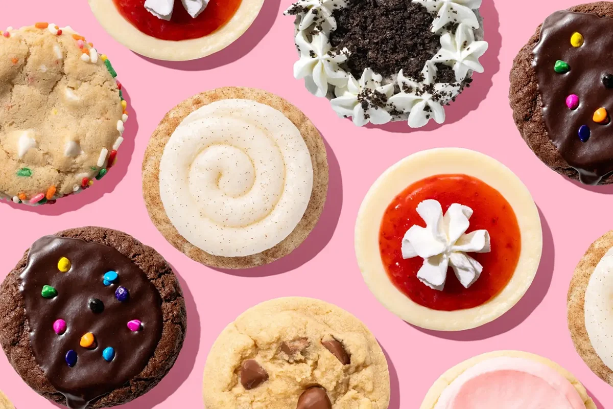 Assorted cookies and mini cheesecakes with frosting, sprinkles, and fruit toppings on a pink background.