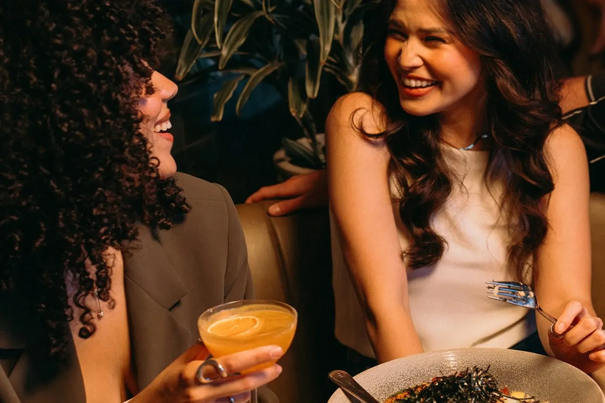 Two women smiling and enjoying a meal and cocktail in a cozy restaurant setting.