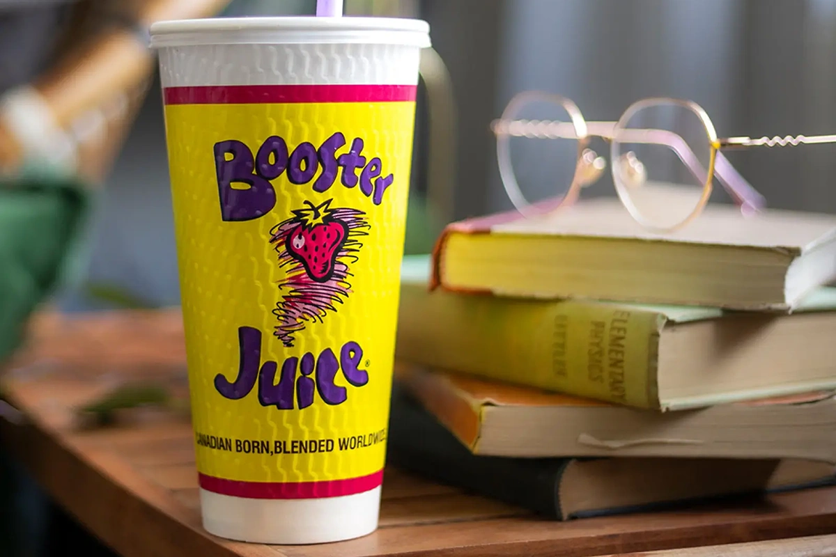Booster Juice branded yellow cup with purple text and a strawberry logo on a wooden table next to stacked books and glasses.