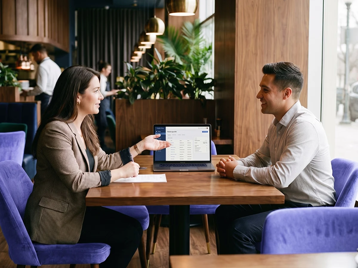Two professionals having a discussion at a wooden table with a laptop displaying data, in a modern office setting.