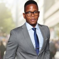 Professional man wearing a gray suit, blue tie, and glasses standing outdoors with blurred city background.