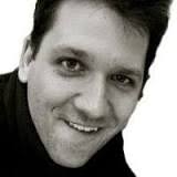 Close-up of a man with short dark hair smiling and looking at the camera against a white background.