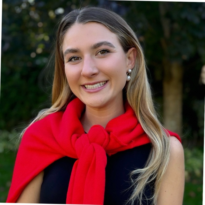 Smiling young woman with long blonde hair wearing pearl earrings and a bright red sweater tied around her shoulders outdoors.