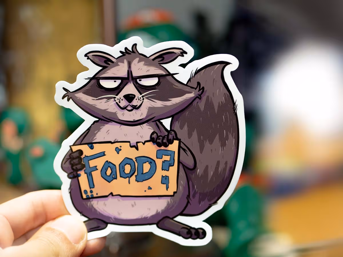 Das Bild zeigt einen Aufkleber, der in einer Hand gehalten wird. Der Aufkleber stellt einen Cartoon-Waschbären dar, der eine Sonnenbrille trägt und ein Schild mit der Aufschrift "FOOD?" hochhält. Der Waschbär hat einen leicht genervten oder müden Gesichtsausdruck, und das Schild ist in einer groben, kindlichen Schriftart geschrieben, mit einigen Farbspritzern rund um die Buchstaben. Der Hintergrund ist unscharf, was darauf hindeutet, dass der Fokus auf dem Aufkleber liegt. Der Waschbär ist überwiegend in Brauntönen gehalten, mit weißen Akzenten um die Augen und dunkleren Farbtönen am Körper.
