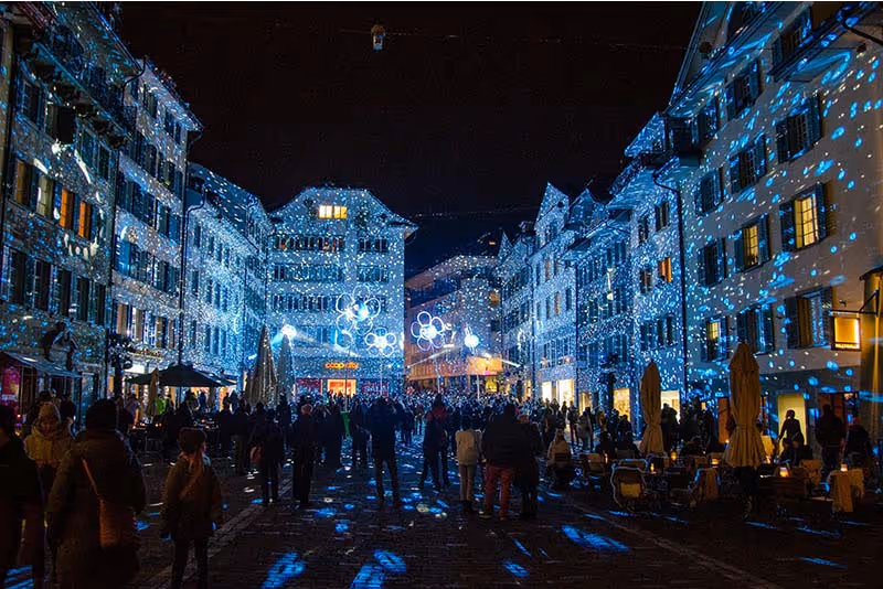 Eine Innenstadt bei einem Kulturevent bei Nacht. SIe ist in blaues Licht gehüllt und auf die Fassaden wird eine Lichtshow projeziert.