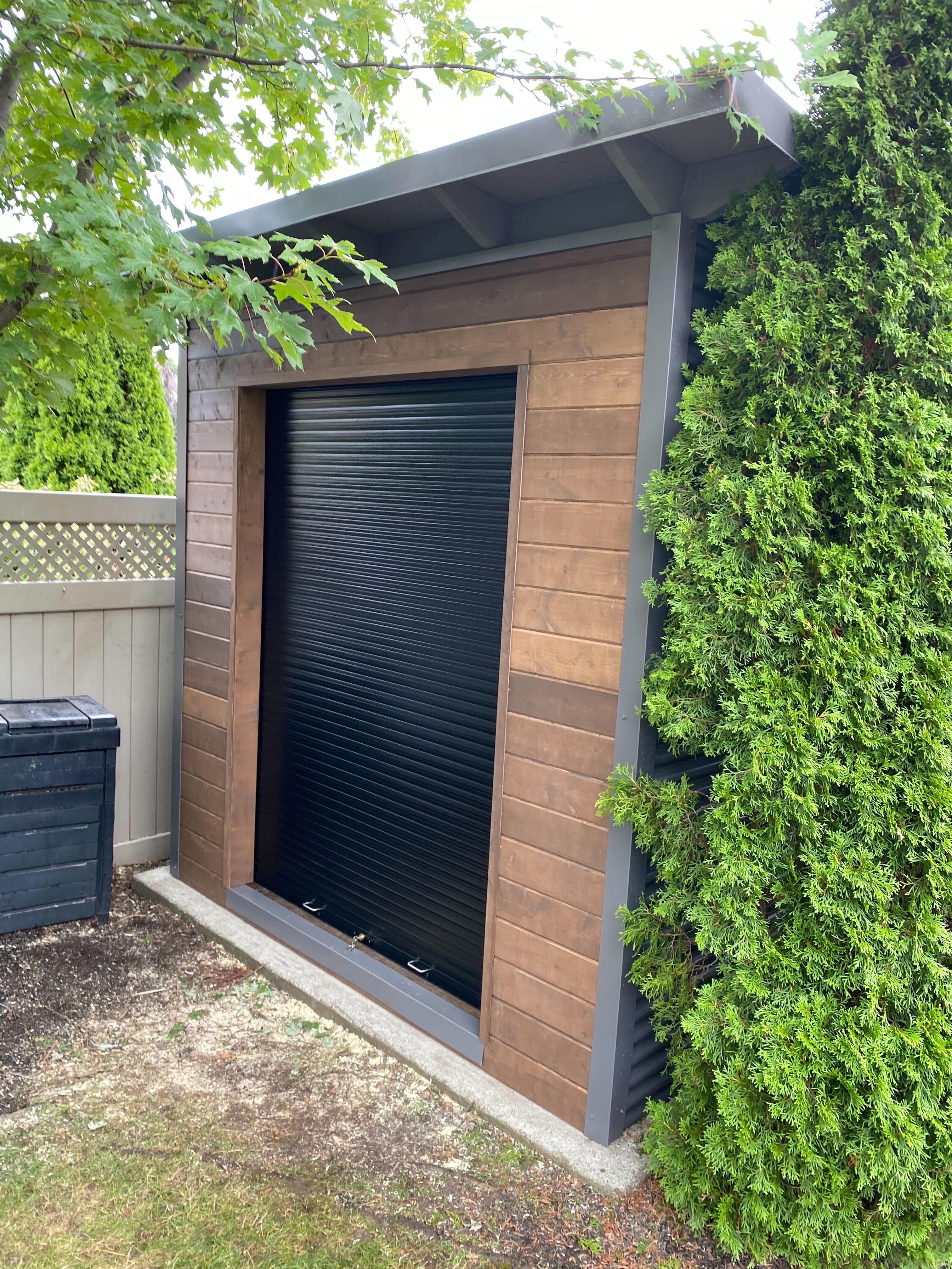 8x8 Essential Shed with charcoal corrugated steel siding, Muskoka Brown pine front wall, and a black aluminum roll-up door installed in Oakville Ontario by Backyard Escape Studios.