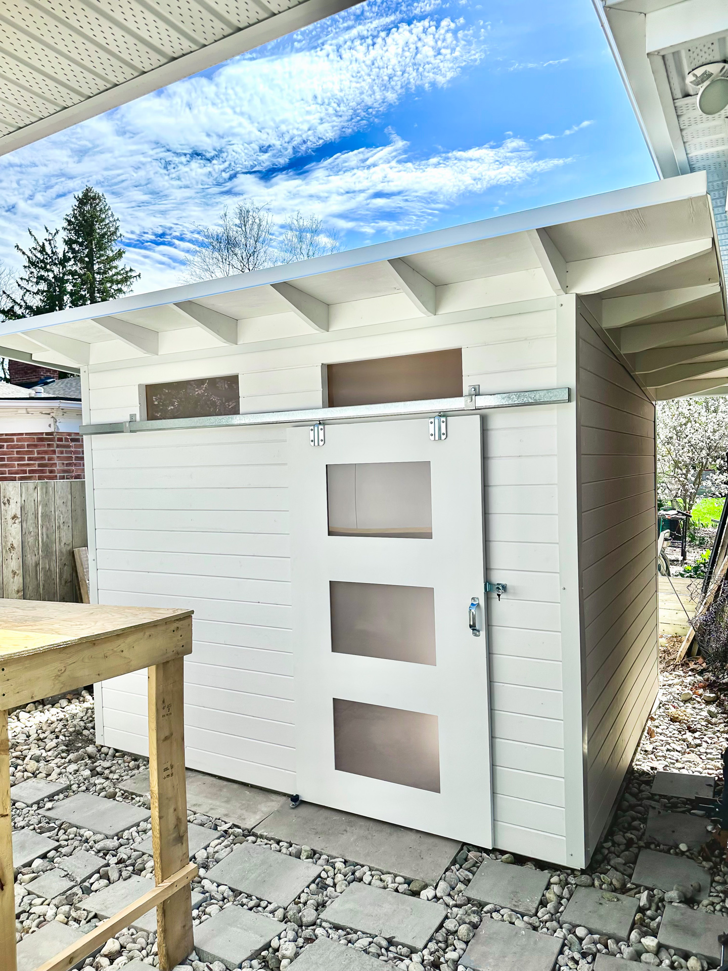 Close-up of 10x8 Advanced Storage Shed front wall in London Ontario showing White Satin sliding barn door with frosted lexan glass panels and transom windows under exposed rafter overhang, installed by Backyard Escape Studios.