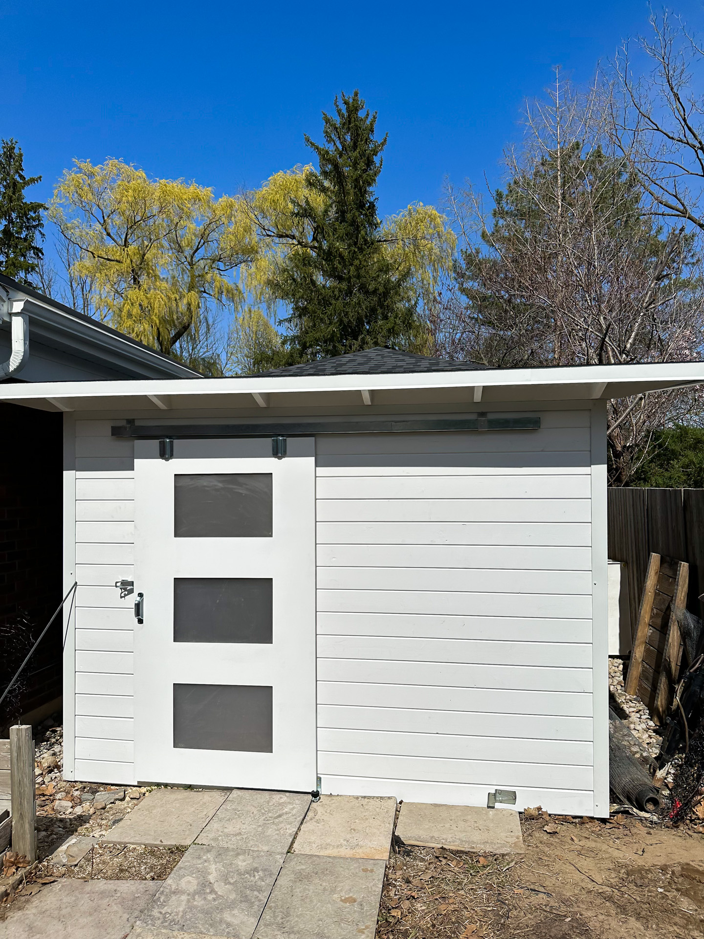 Front elevation of 10x8 Advanced Storage Shed in London Ontario with White Satin horizontal pine T+G siding, sliding barn door, and exposed rafter soffits against a clear blue sky, installed by Backyard Escape Studios.