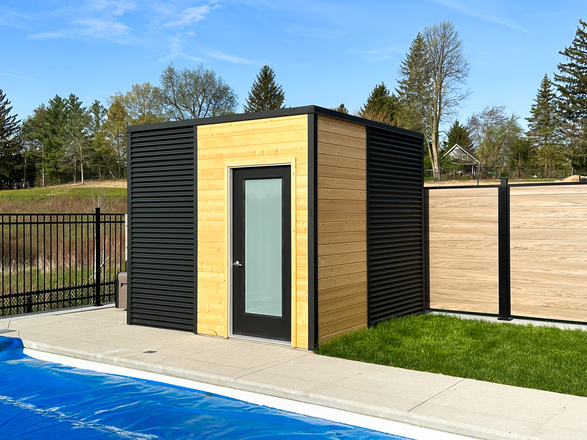 10x9.5 Cube Shed pool shed with split jet black corrugated steel and raw pine tongue-and-groove walls, frosted glass inswing door, installed poolside in Kitchener Ontario by Backyard Escape Studios.