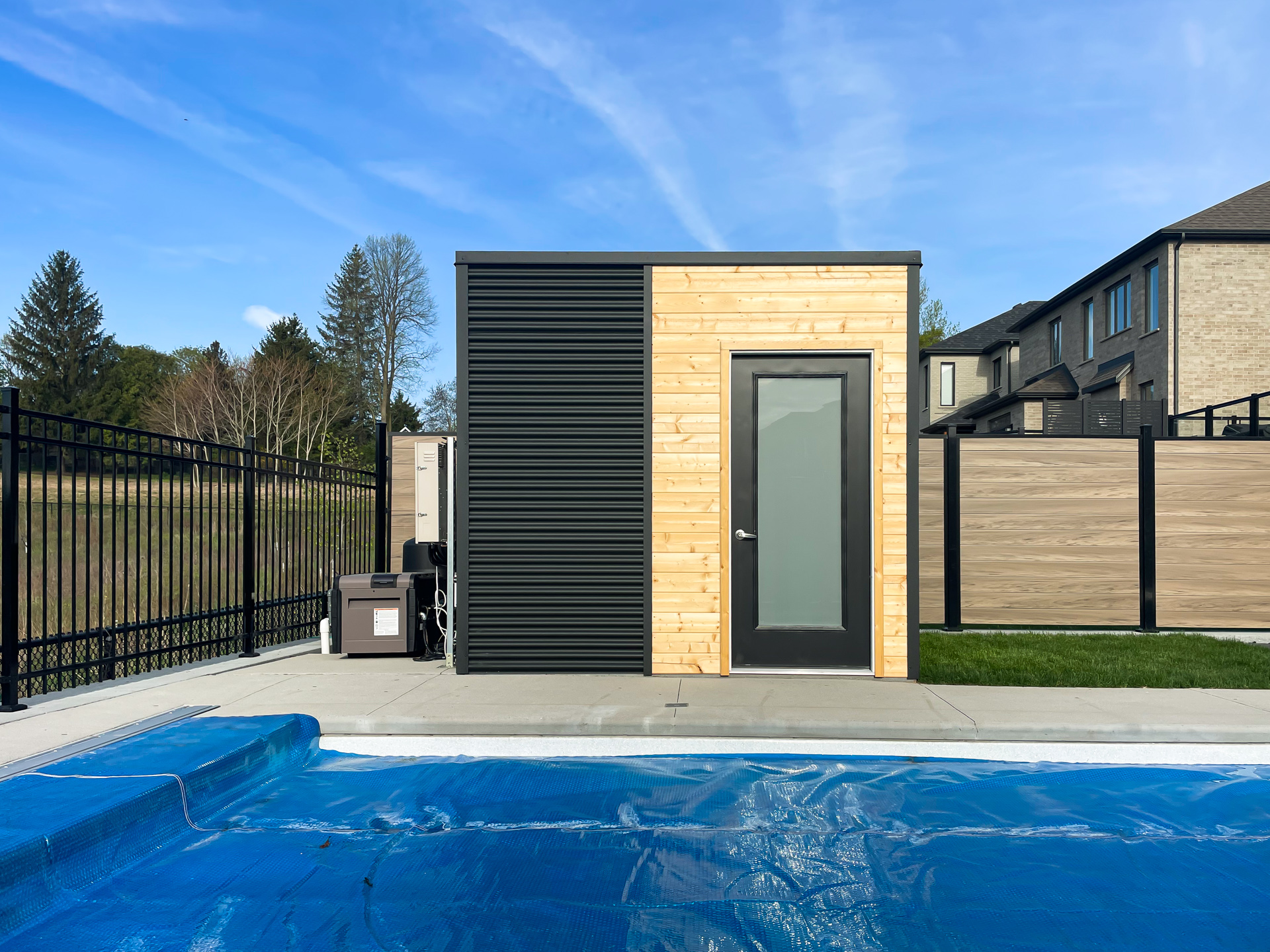 Three-quarter view of a 10x9.5 Cube Shed pool shed showing jet black corrugated steel right wall and pine front panel, installed poolside in Kitchener Ontario by Backyard Escape Studios.