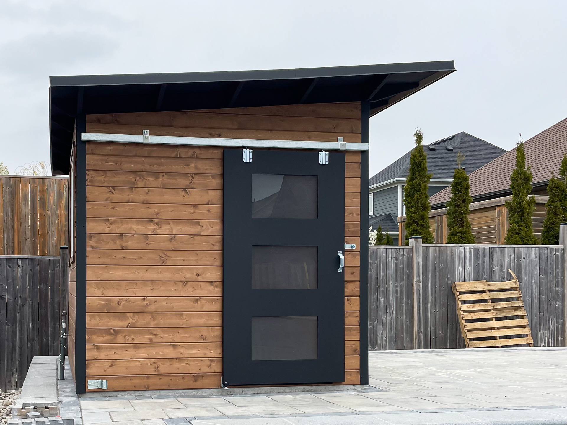 10x8 Advanced Shed with cedar tongue-and-groove siding in Muskoka Brown stain and a sliding barn door in Black Beauty, installed beside an inground pool in Strathroy Ontario by Backyard Escape Studios.