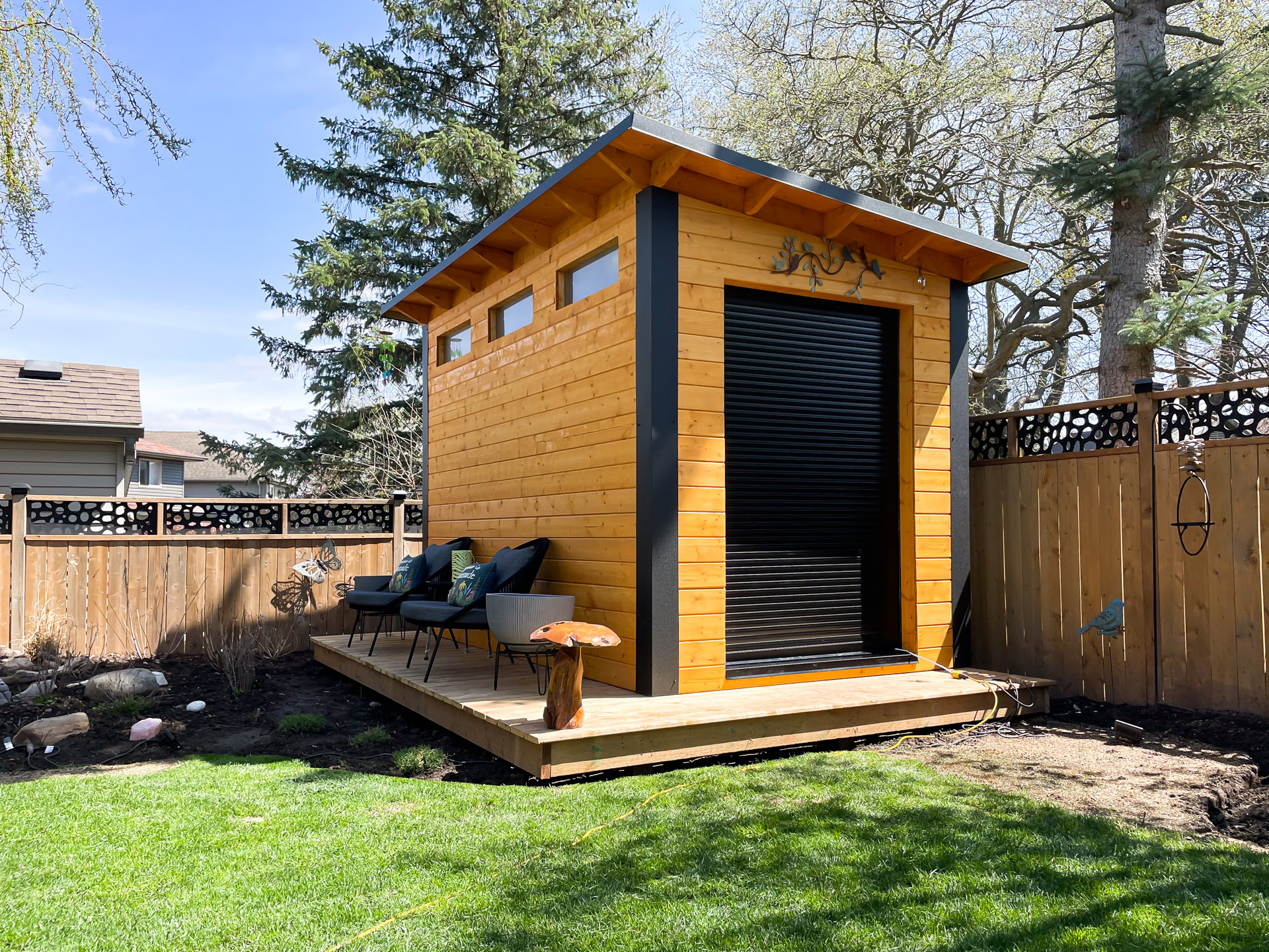 12x7 Essential Shed with cedar-stained pine siding, jet black corrugated steel walls, and aluminum roll-up door installed on a timber deck in London Ontario, installed by Backyard Escape Studios.