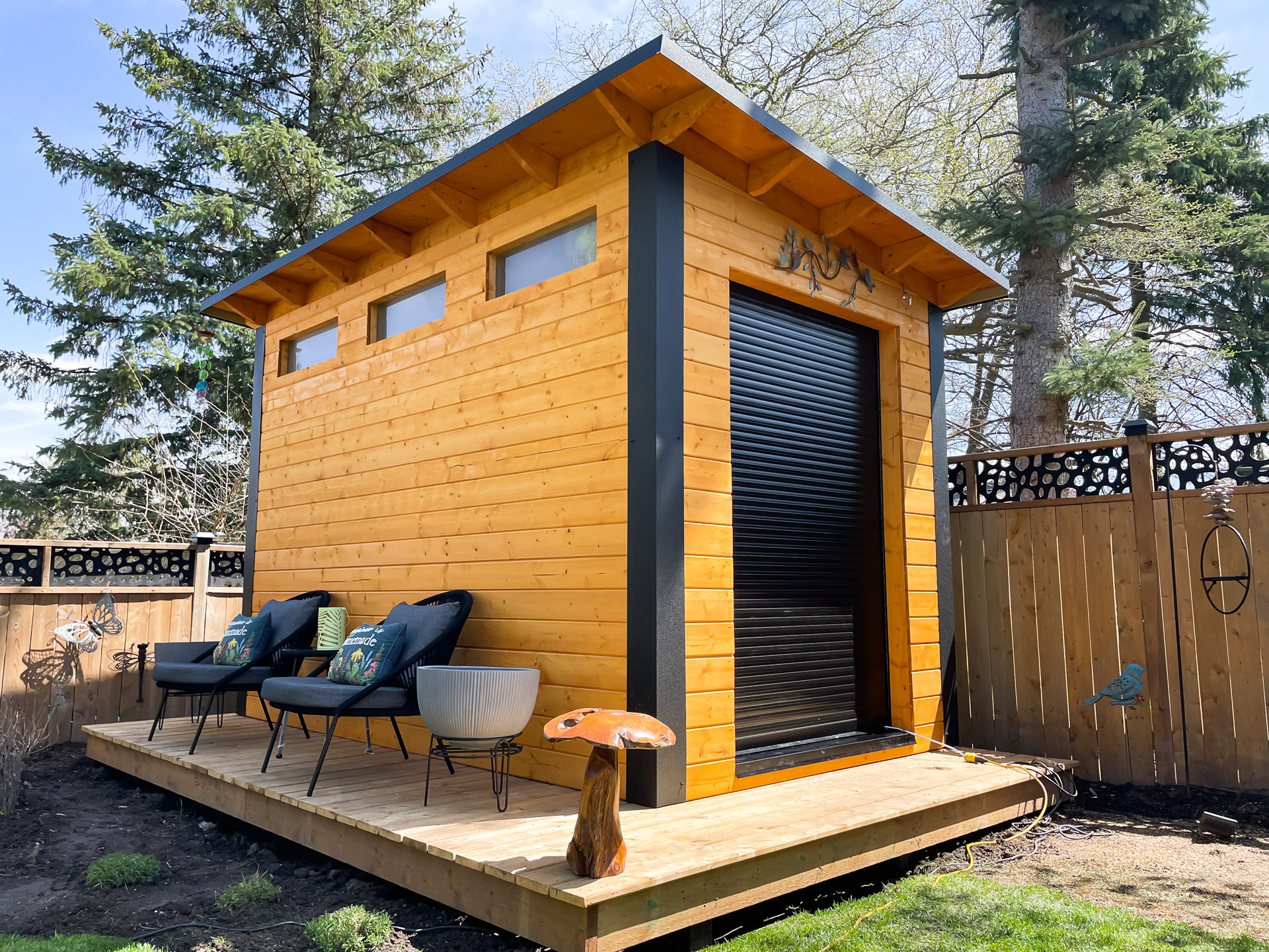 Front view of 12x7 Essential Shed in London Ontario showing three horizontal transom windows and full-width black aluminum roll-up shutter door with cedar-stained pine siding, installed by Backyard Escape Studios.