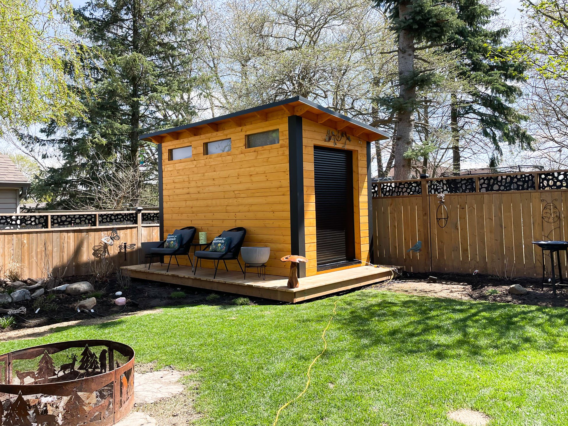 12x7 Essential Shed in London Ontario with jet black corrugated steel back wall and cedar-stained pine front, viewed from the lawn with fire pit in foreground, installed by Backyard Escape Studios.