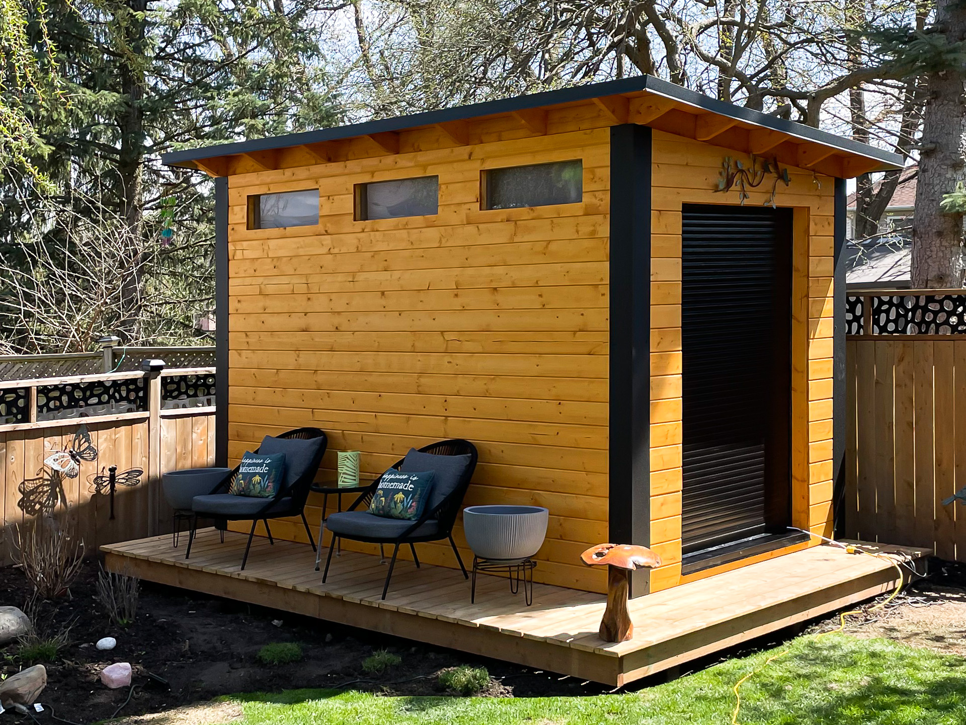 12x7 Essential Shed with cedar-stained pine siding and black aluminum roll-up door on a timber deck in London Ontario, installed by Backyard Escape Studios.
