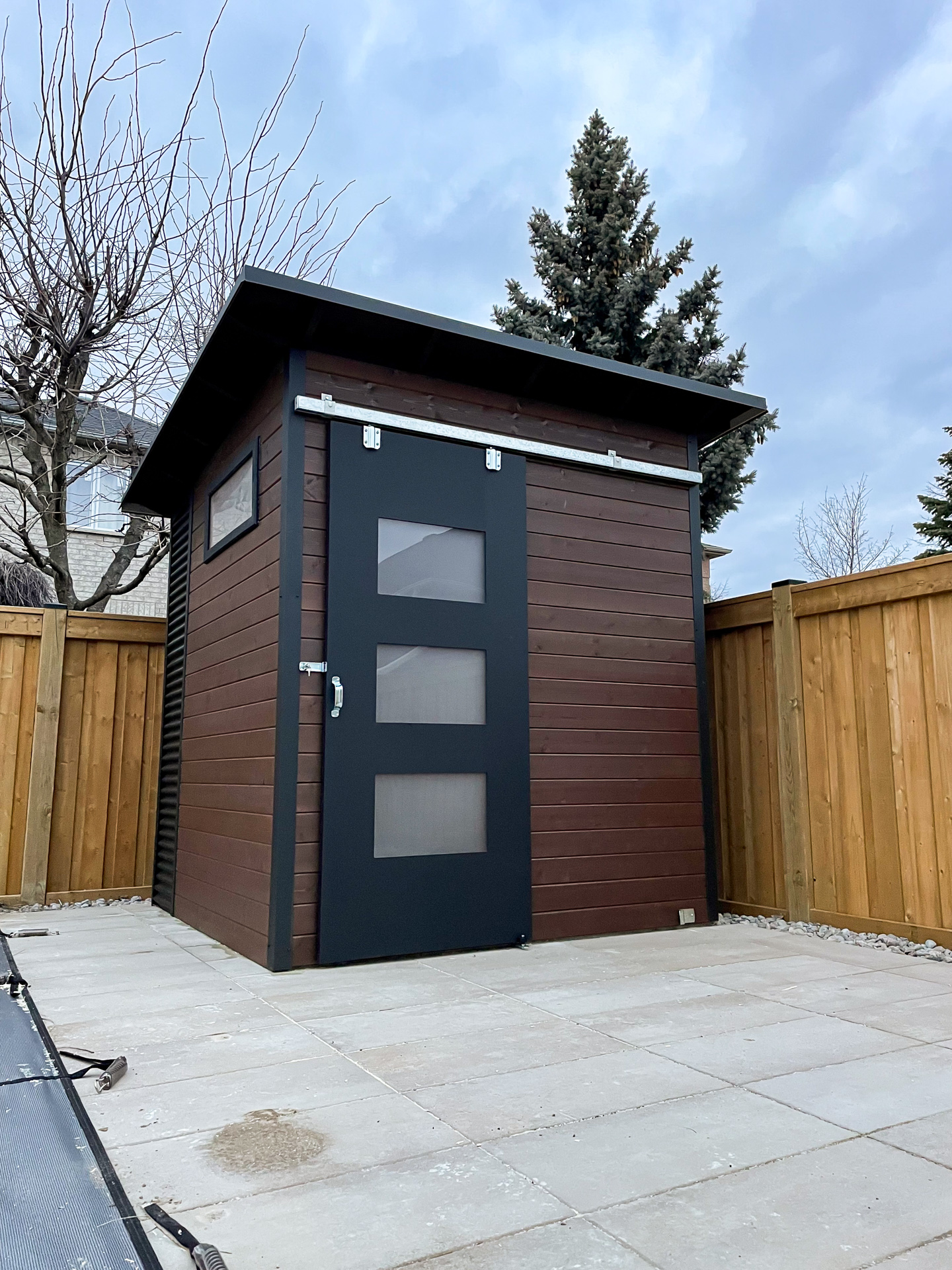 8x8 Essential Shed with Jet Black corrugated steel siding, SICO Walnut Brown stained pine front wall, and a sliding barn door with frosted Lexan glass panels in Vaughan Ontario, installed by Backyard Escape Studios.