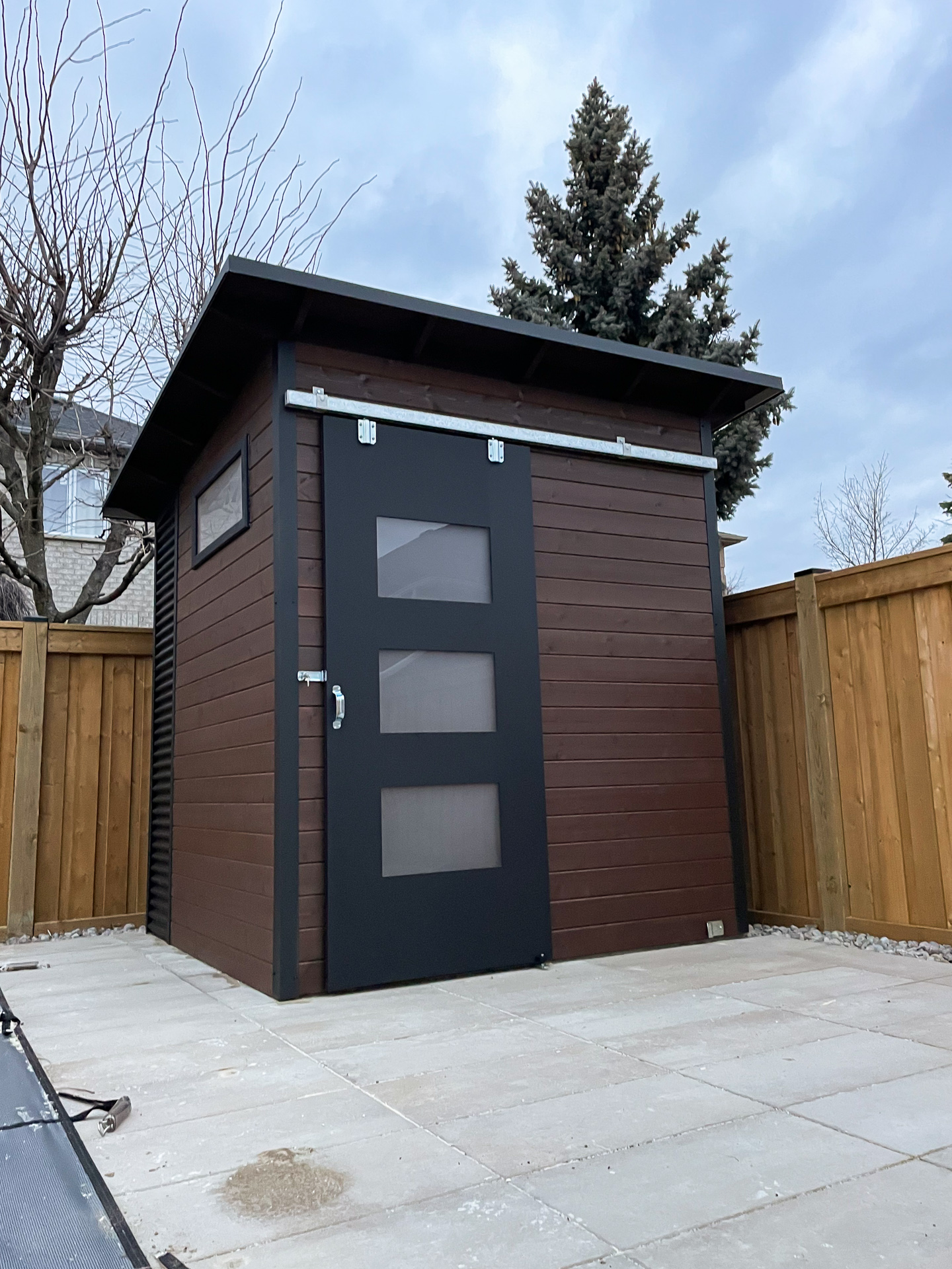 8x8 Essential Shed in Maple Ontario with Walnut Brown pine front wall, Jet Black corrugated steel siding, and a sliding barn door with frosted Lexan glass windows installed by Backyard Escape Studios.