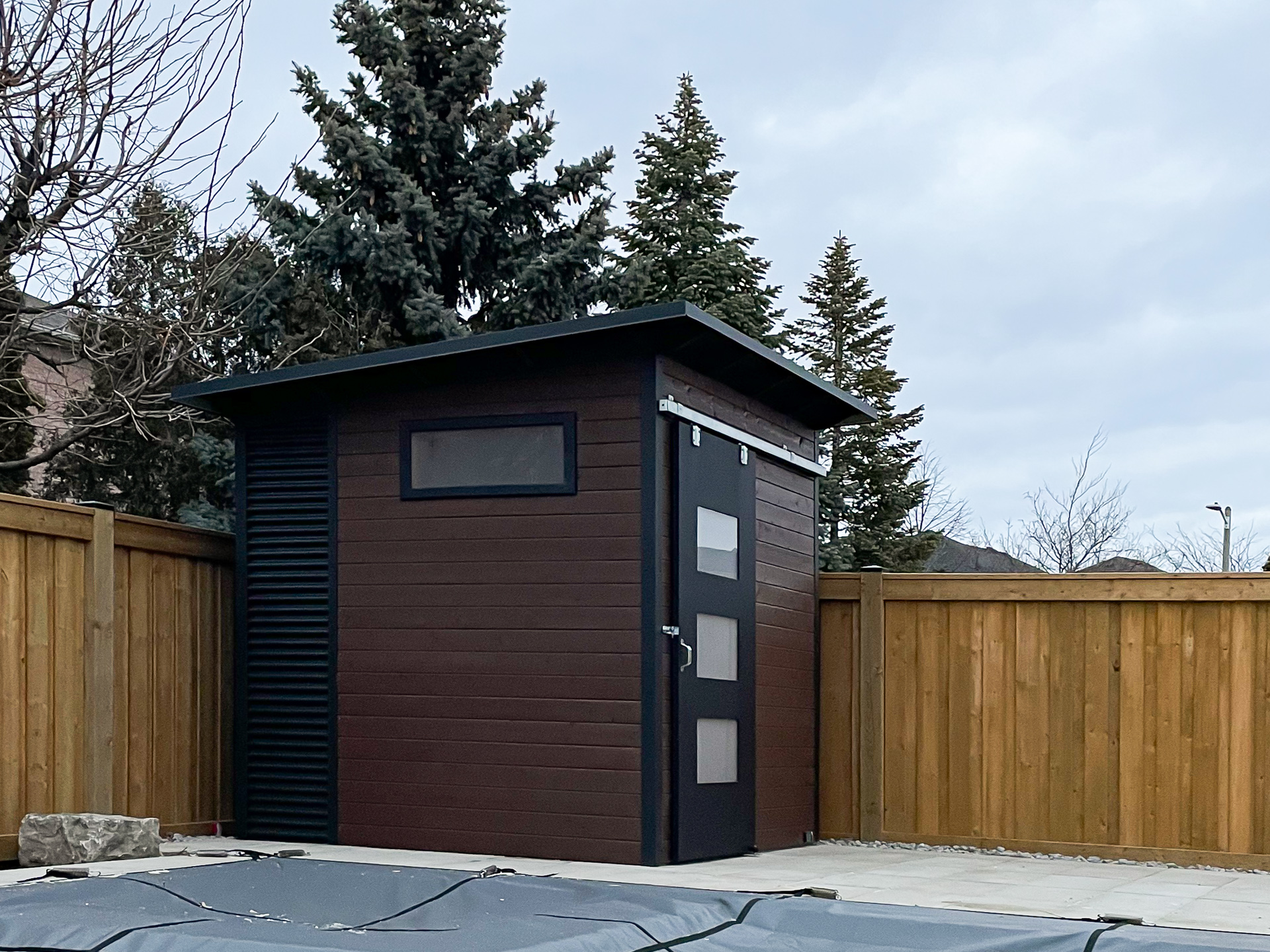 8x8-pool-shed-maple-ontario-jet-black-corrugated-steel-side.jpg Alt: Side and rear view of an 8x8 Essential Shed in Maple Ontario clad in Jet Black corrugated steel with a frosted transom window and walnut-stained pine accent wall, installed by Backyard Es