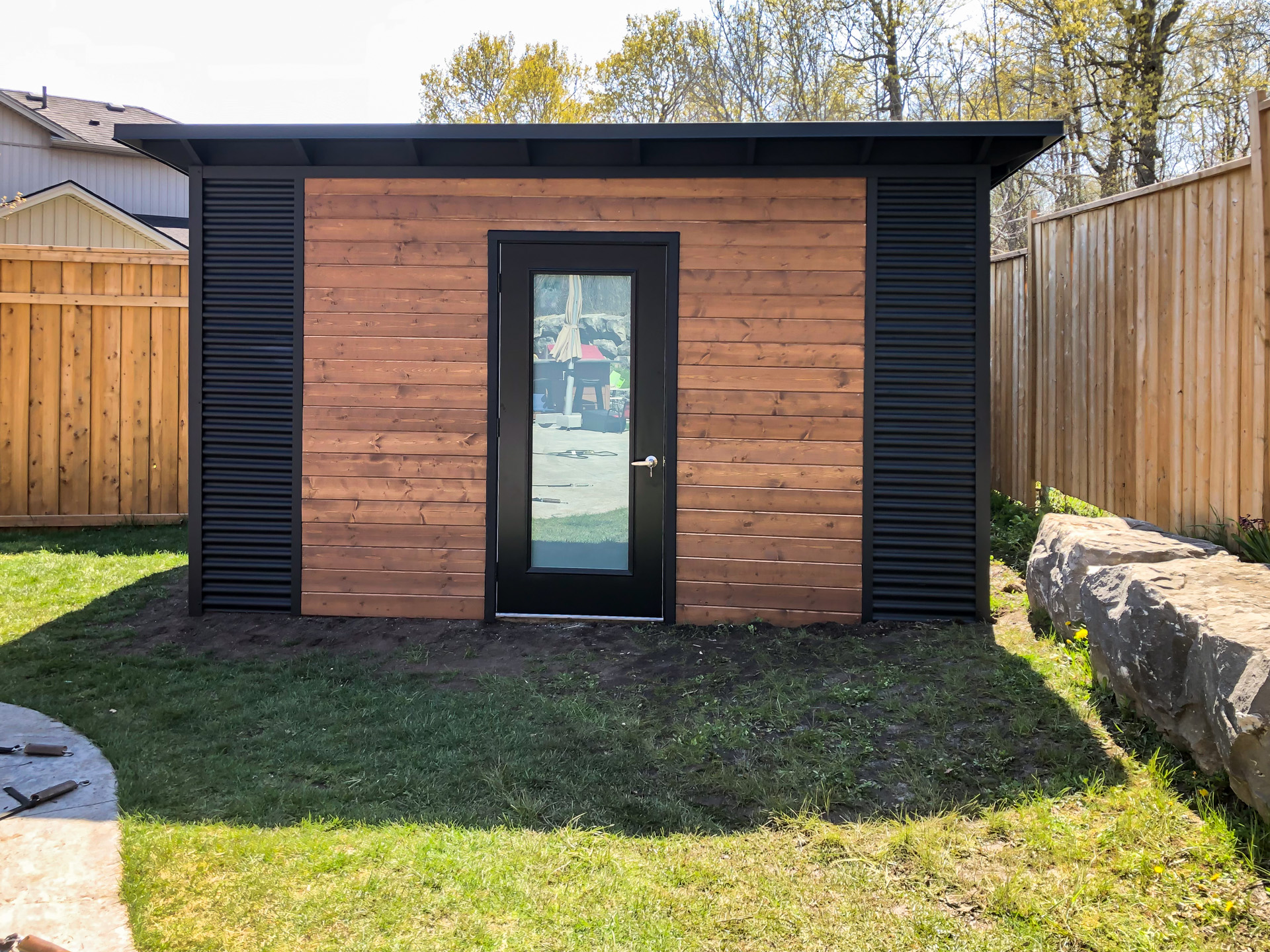 Front view of a 14x10 Essential Shed in London Ontario with Muskoka Brown horizontal pine panelling, Jet Black corrugated steel corners, and a full-glass frosted outswing door, installed by Backyard Escape Studios.