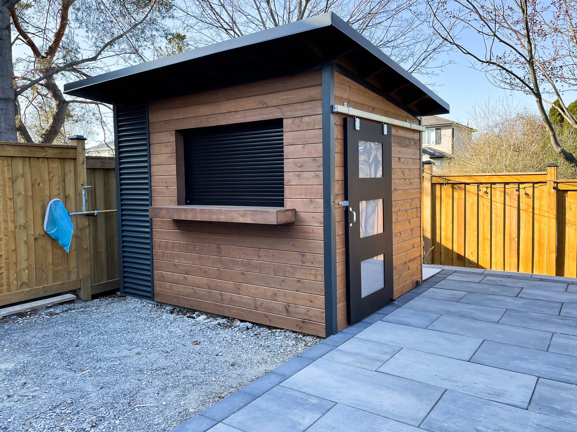 10x8 Essential pool shed with sliding barn door with frosted Lexan glass panels and roll-up bar window on stained pine front wall in North York Ontario, installed by Backyard Escape Studios.