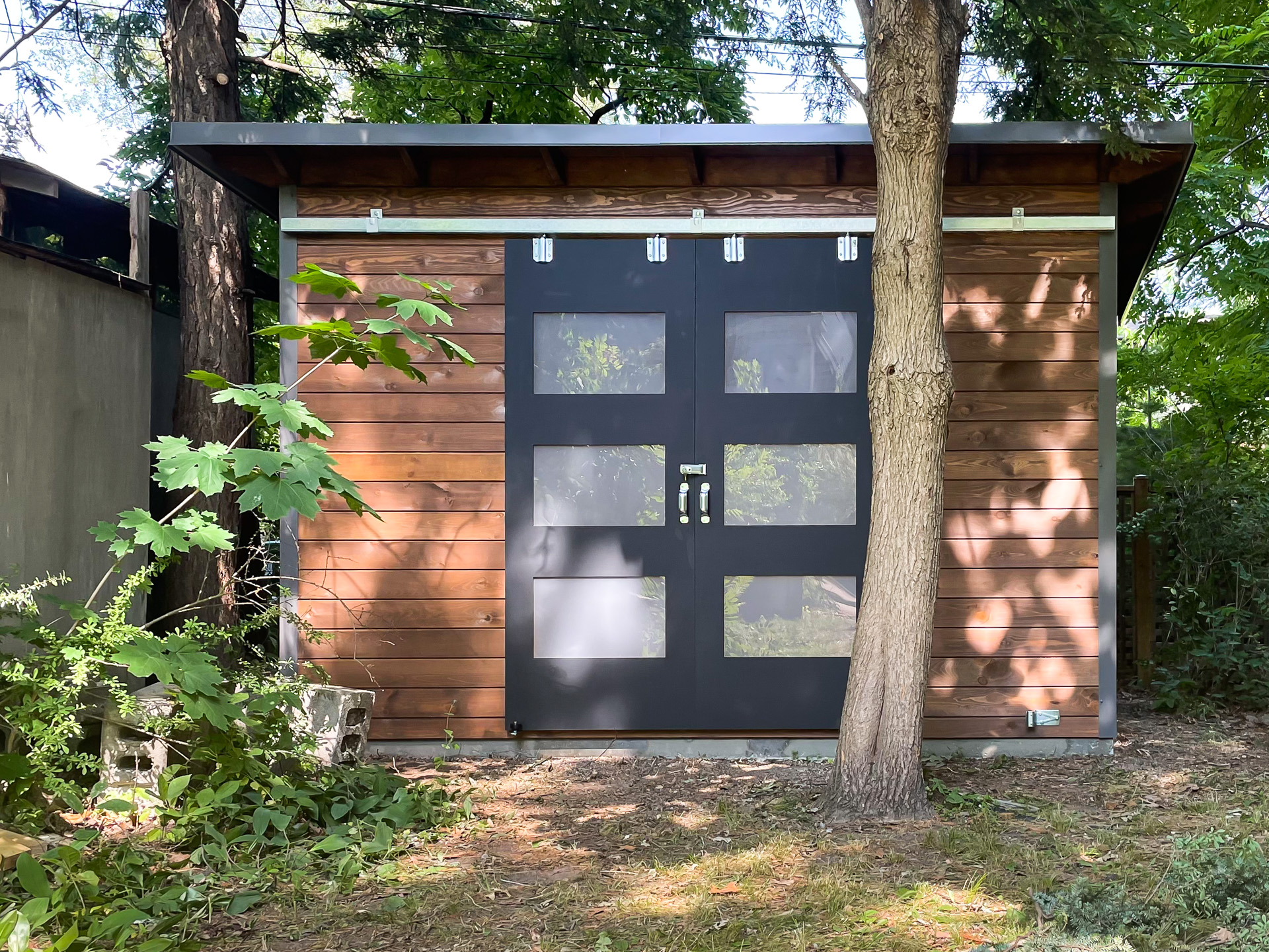 Front view of a 12x8 Essential Shed in Toronto Ontario showing double sliding barn doors with frosted Lexan glass panels and Muskoka Brown stained pine T+G siding, installed by Backyard Escape Studios.