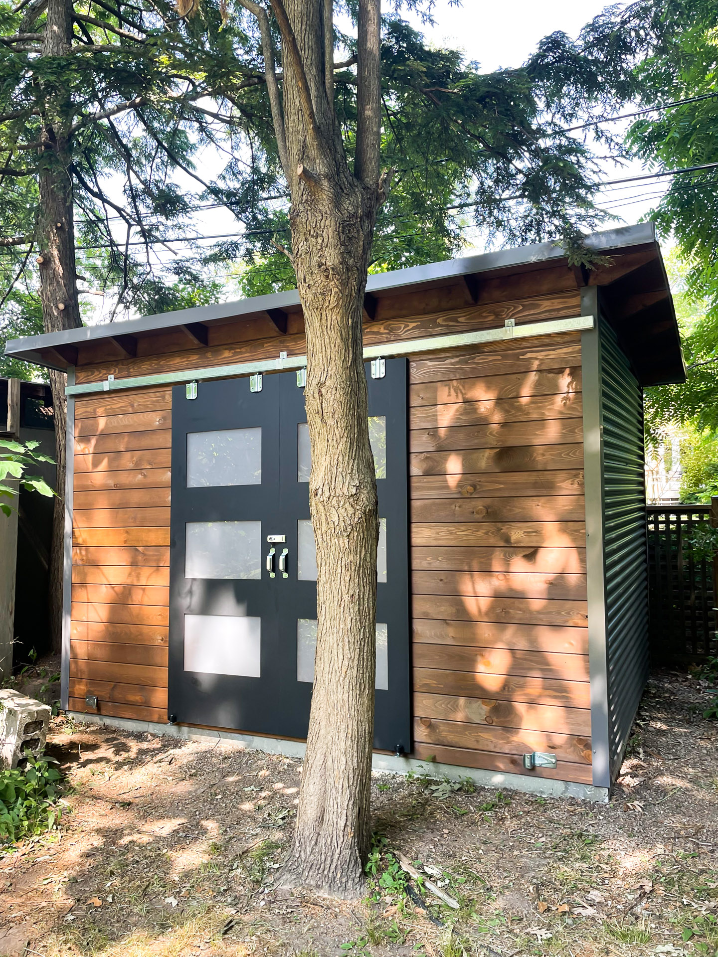 Side-angle view of a 12x8 Essential Shed in Toronto Ontario with charcoal corrugated steel siding and Muskoka Brown pine front wall with double sliding barn doors, installed by Backyard Escape Studios.